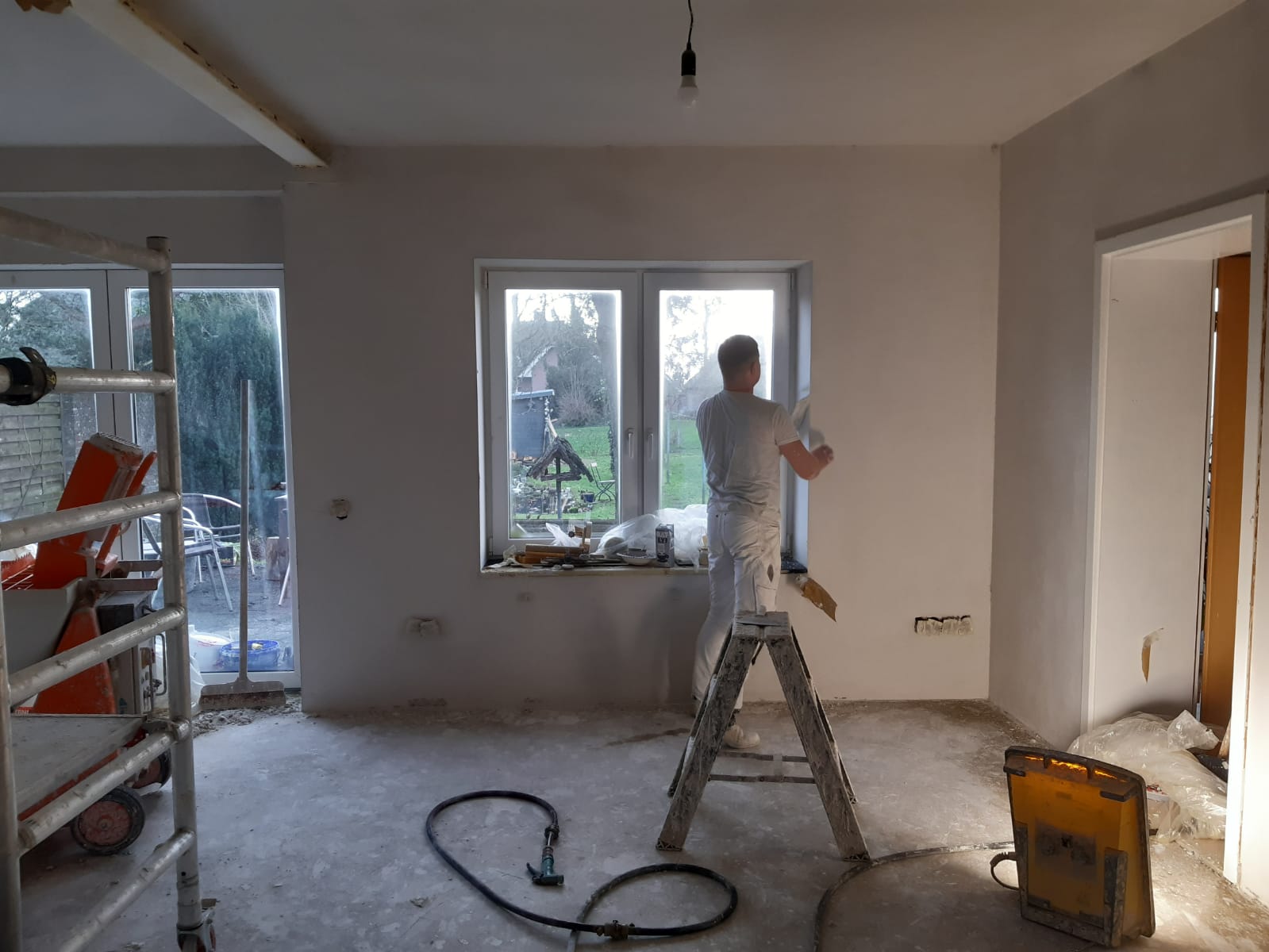 Alex aan het werk op een trapje bij het raam, met steiger en stucmachine in de kamer